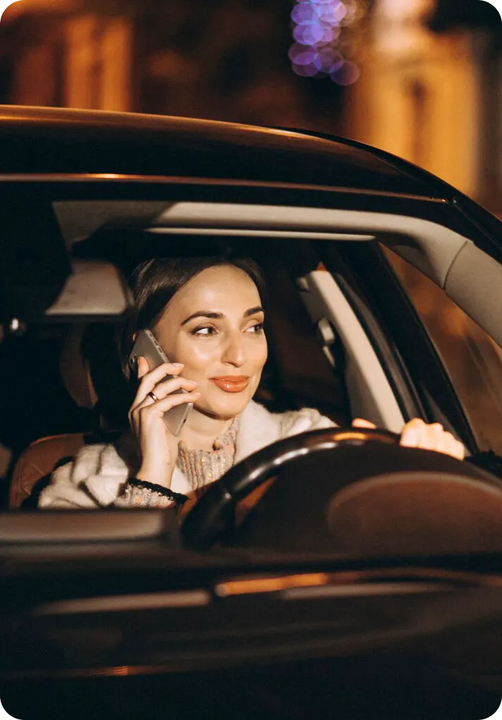 A woman in a navy hijab sits in a car, holding a phone to her ear. She looks thoughtful, gazing outside. The interior is neutral-toned, creating a calm mood.